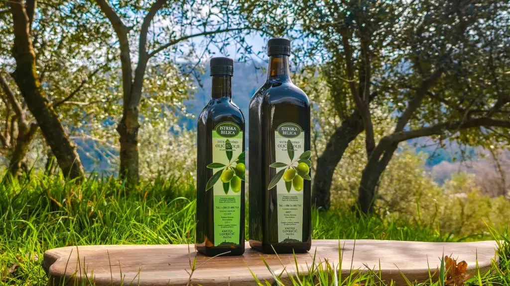 Dve steklenici ekstra deviškega oljčnega olja Kmetije Lovrečič (Istrska belica, 0,75 L in 1 L) na leseni mizi v obsijanem oljčniku.